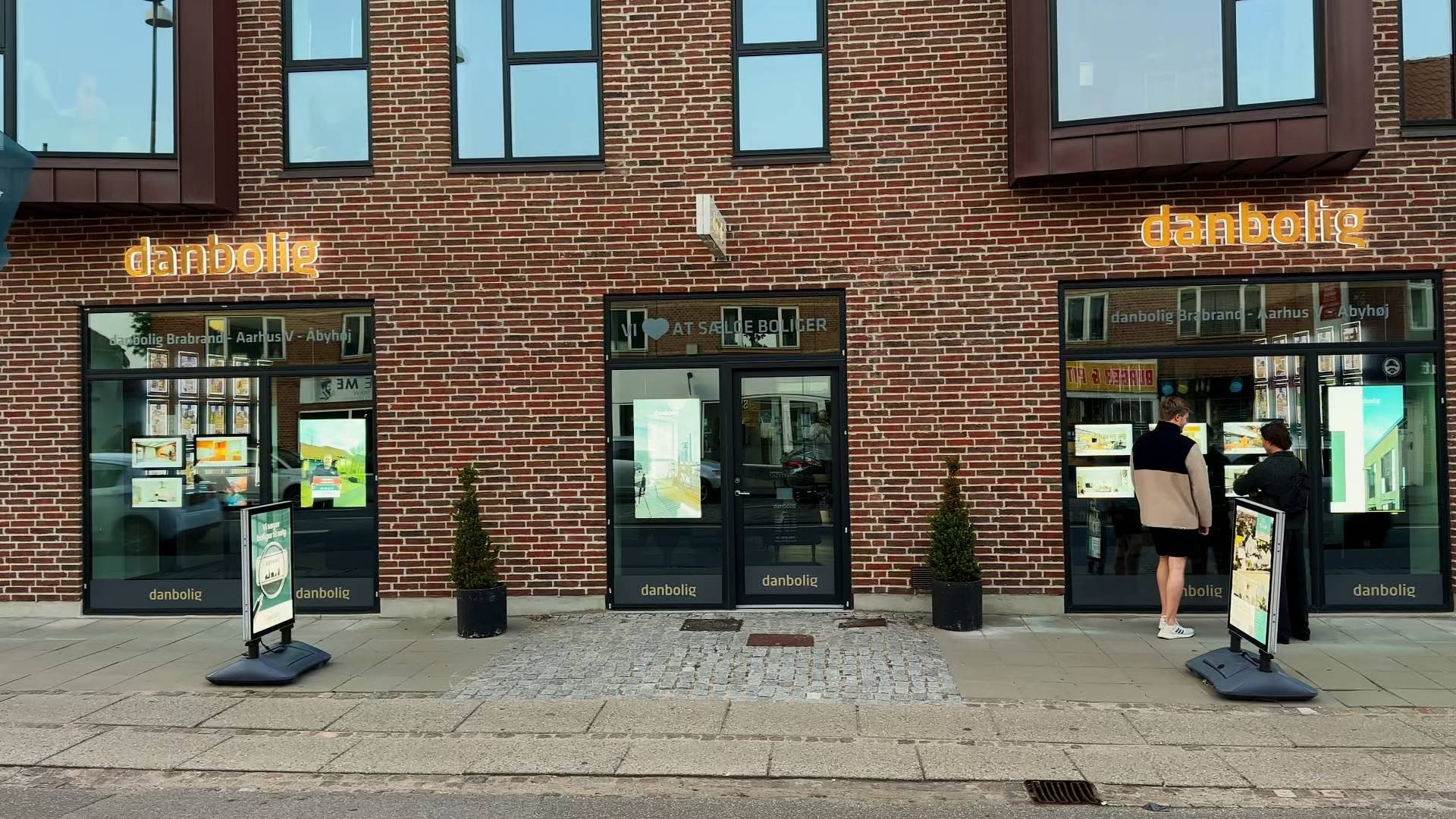 danbolig Åbyhøj i røde mursten: digitale skærme lyser i begge vinduespartier, og der står fortovsskilte foran indgangen.