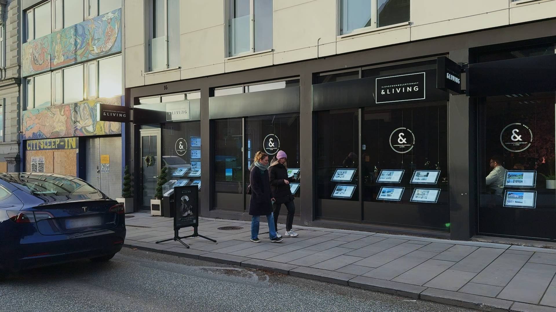 &Living i Aarhus set fra fortovet: flere stående skærme i vinduet viser boliger, mens to forbipasserende standser og kigger.
