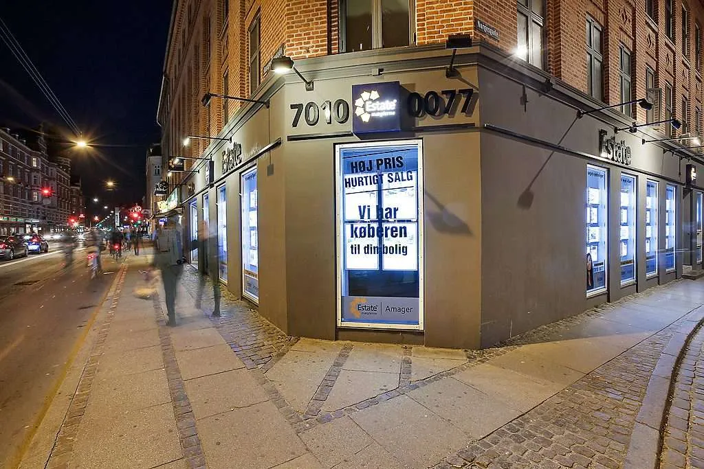 Estate Amagers hjørnebutik i skumringen med en stor belyst plakat i vinduet med teksten Høj pris, hurtigt salg — vi har køberen til din bolig.