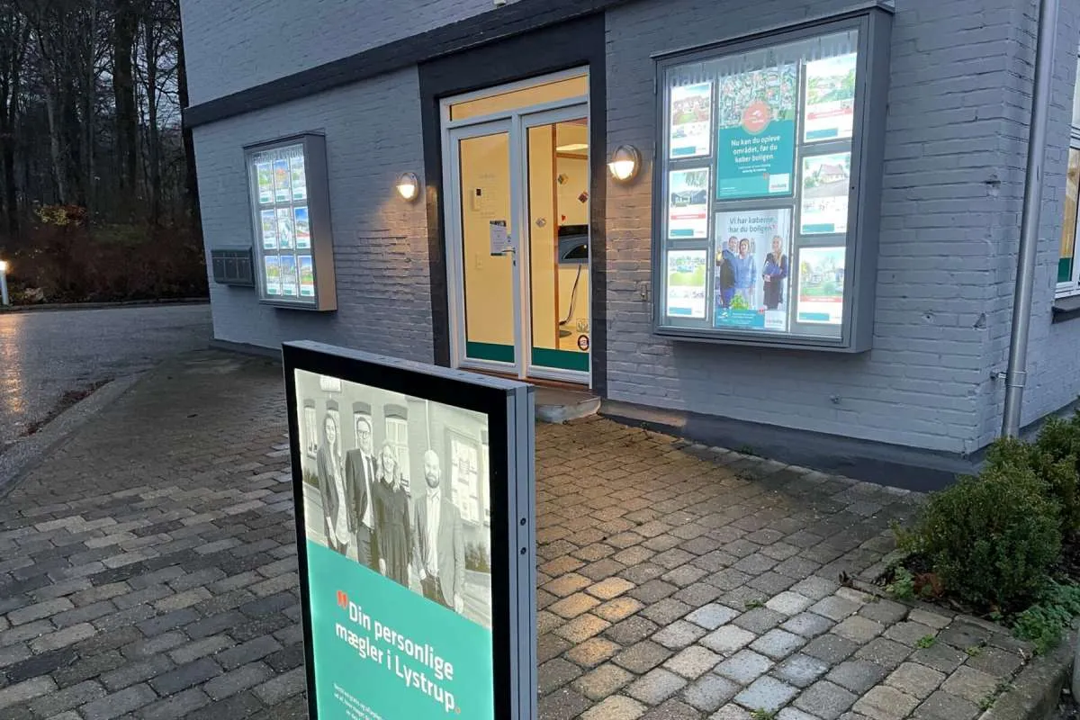 Homes butik i Lystrup ved aftenstid med belyste plakatholdere i to vinduer og en fritstående lysende stander på fortovet.